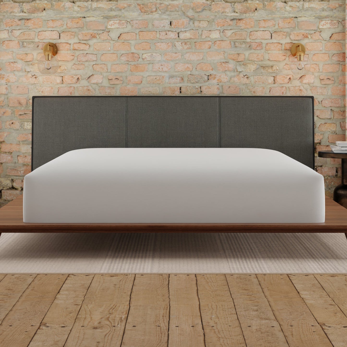 Bed with gray upholstered headboard and white mattress against a brick wall. White mattress is covered with the Essentials mattress protector
