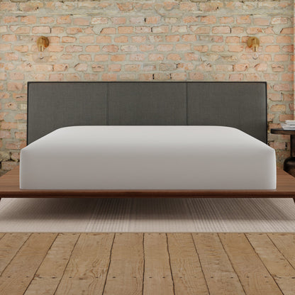 Bed with gray upholstered headboard and white mattress against a brick wall. White mattress is covered with the Essentials mattress protector