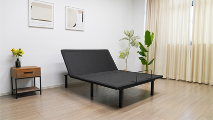 Black adjustable bed in a room with a nightstand, plant, and curtains. The head of the adjustable bed base is elevated.