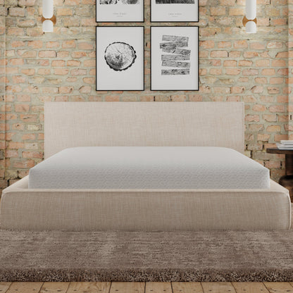 upholstered Bed with a mattress covered with Snow mattress protector. Bed is against a brick wall with framed artwork.