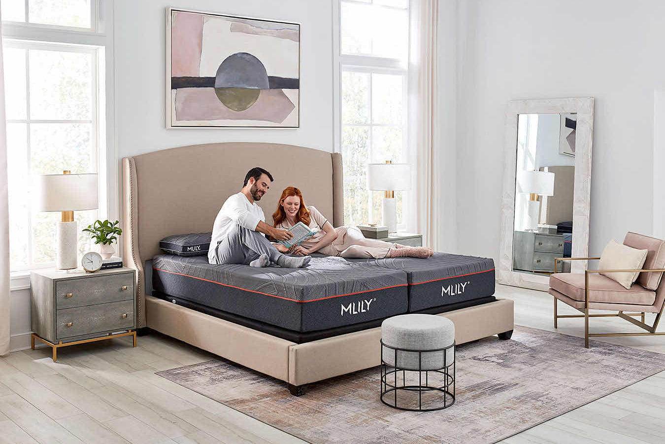 A couple relaxes on a dark gray Mlily mattress on a beige upholstered bed in a bright, modern bedroom with light wood floors, a nightstand, and a chair.