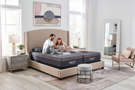A couple relaxes on a dark gray Mlily mattress on a beige upholstered bed in a bright, modern bedroom with light wood floors, a nightstand, and a chair.