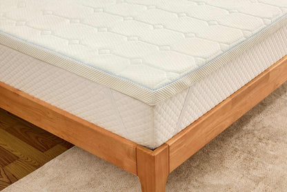 A close-up of a white mattress topper or mattress corner resting on a light wood bed frame over a beige carpet.